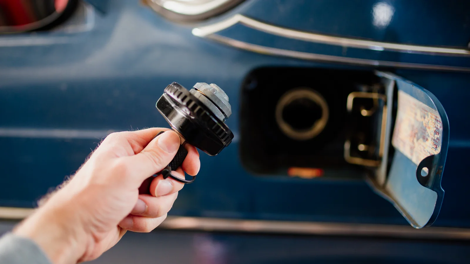 A hand holding a fuel cap beside the open filler of a blue car